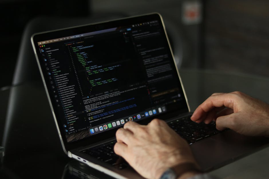 A pair of hands typing on a sleek, modern laptop displaying lines of code on a dark background, with the laptop placed on a dark surface. The scene suggests a focus on programming or digital work, wit